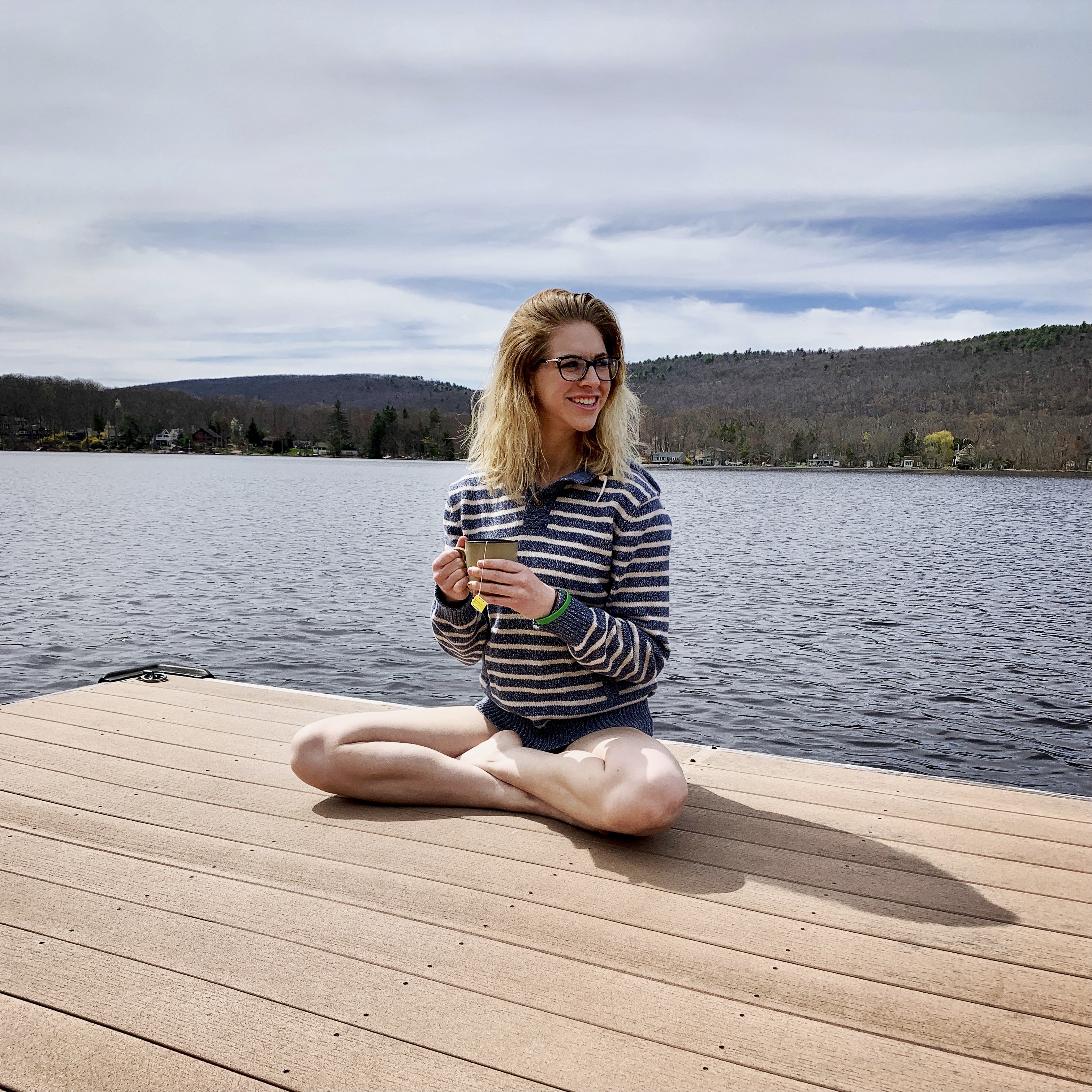 girl sitting on a dock with a lake behind her, holding a cup of tea