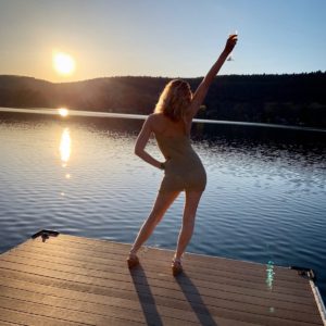 blonde birthday girl holding up a glass to cheers the sunset, looking out over a lake, standing on a dock