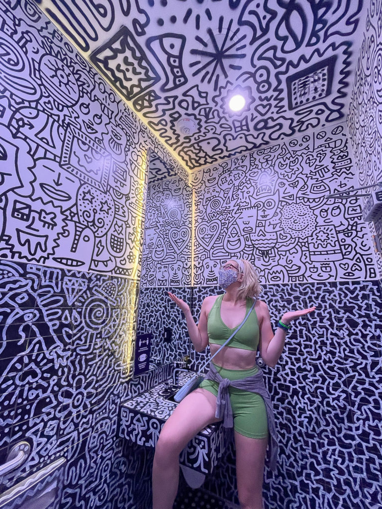 blonde girl with a blue mask and green leggings set sitting on a counter in a massively decorated bathroom.  The walls are totally covered with black and white drawings