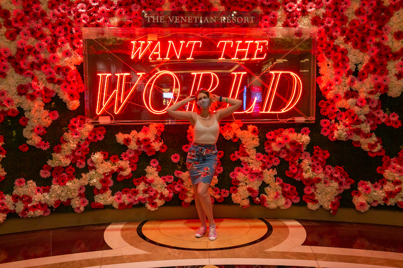 blonde girl in an outfit, packed for her Vegas trip, in front of a wall of pretty pink flowers and a neon sign
