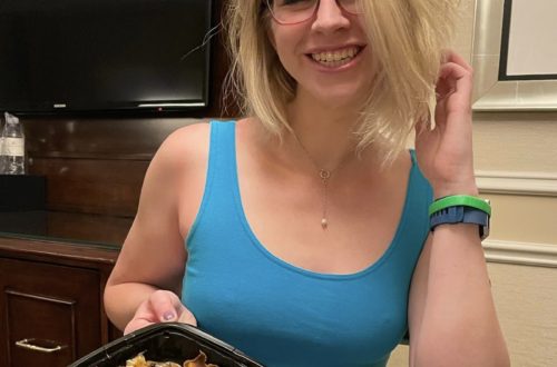blonde girl in a blue top smiling at the camera and holding up a black to go container of food- mashed potatoes and chicken in the Venetian resort (Las Vegas)
