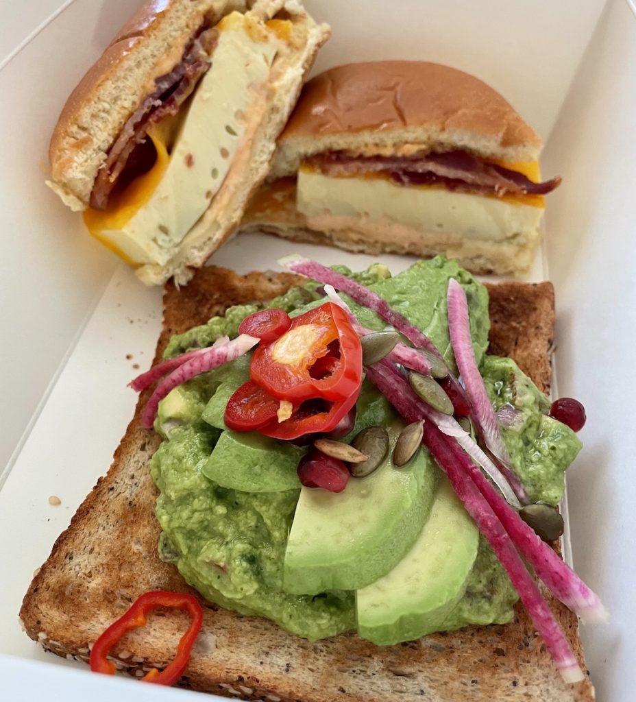 Avocado toast with onions and chilis in a white box alongside a breakfast sandwich filled with cheese bacon and egg.  Takeout breakfast from Chica in the Venetian Vegas