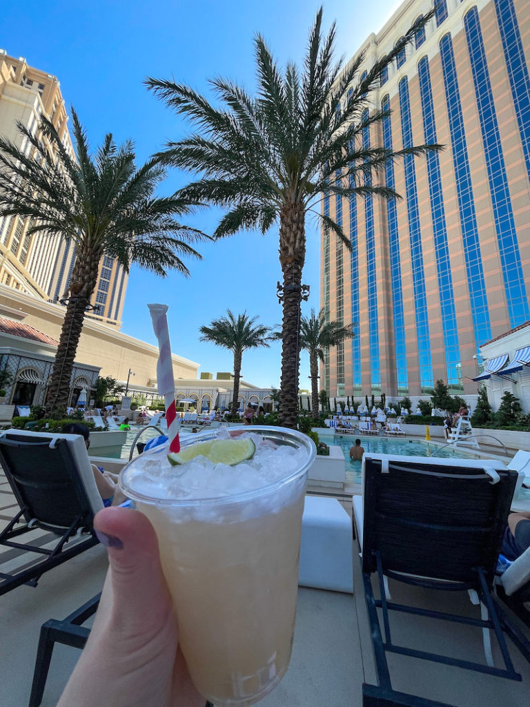 a hand holding a plastic cup filled with an icy mocktail in front of a pool with a tall hotel behind it
