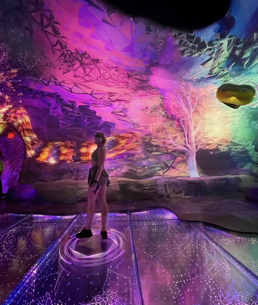 blonde girl standing in the center of the projected desert in OMEGA MART in Las Vegas, purple colored trippy projections on the walls and floor