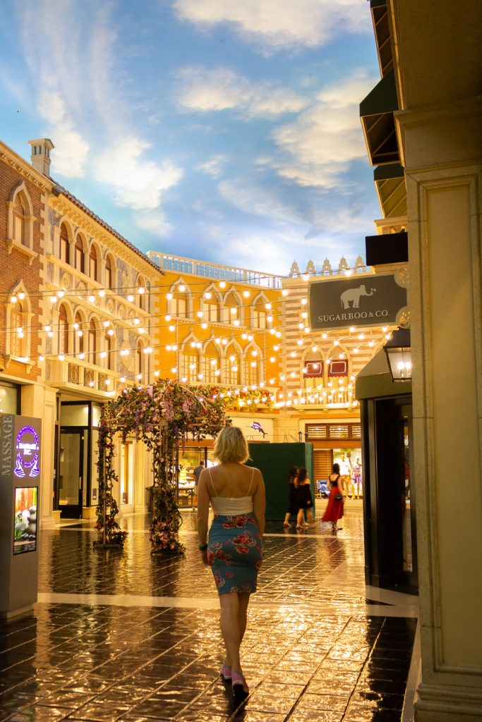 blonde girl with short hair wearing a white camisole and blue flowered skirt, facing away from camera, twinkly lights overhead