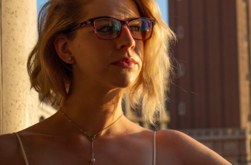 blonde girl in a white tank top and glasses looking off into the distance, backlit by the sun