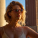 blonde girl in a white tank top and glasses looking off into the distance, backlit by the sun