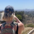 blonde girl with short hair wearing a baseball cap and a colorful backpack, looking away from the camera at the horizon