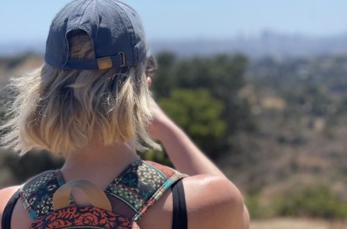 blonde girl with short hair wearing a baseball cap and a colorful backpack, looking away from the camera at the horizon