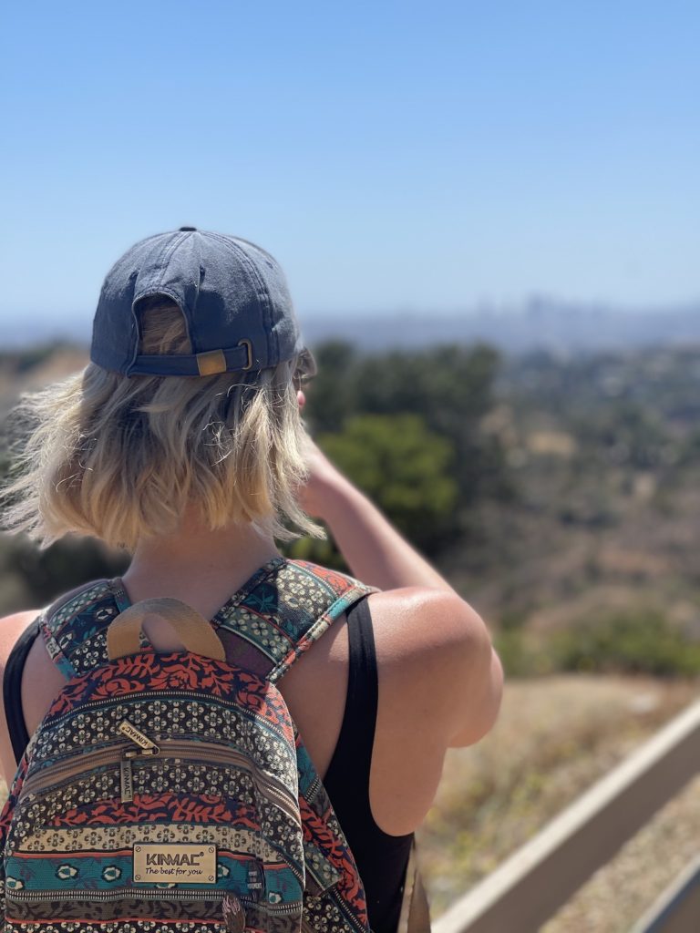 blonde girl with short hair wearing a baseball cap and a colorful backpack, looking away from the camera at the horizon