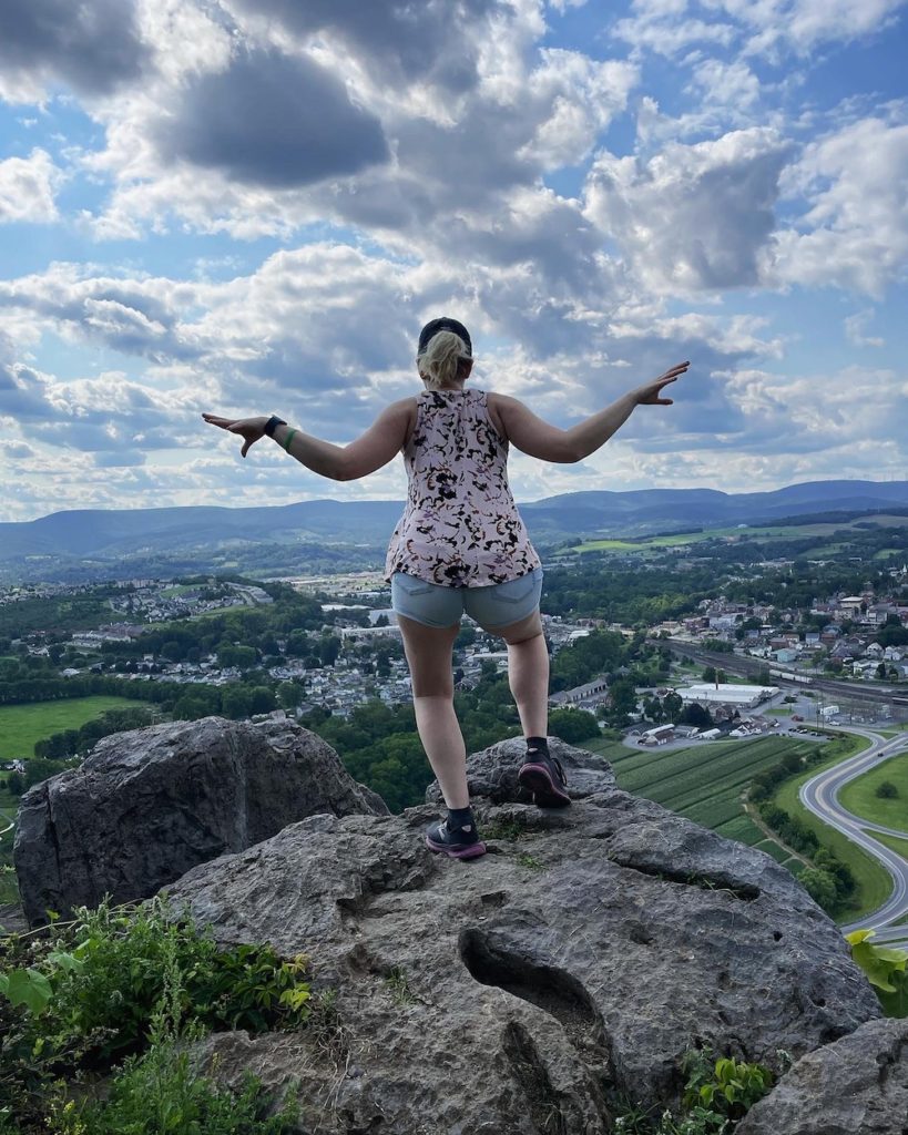 girl with short hair wearing a baseball cap looking away from the camera and over a cliff in PA, arms are up and she is posed like a dragon about to take flight