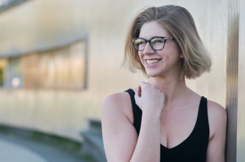 blonde girl with short hair and glasses, wearing a black tank top, looking to the side and smiling in front of an exterior wall