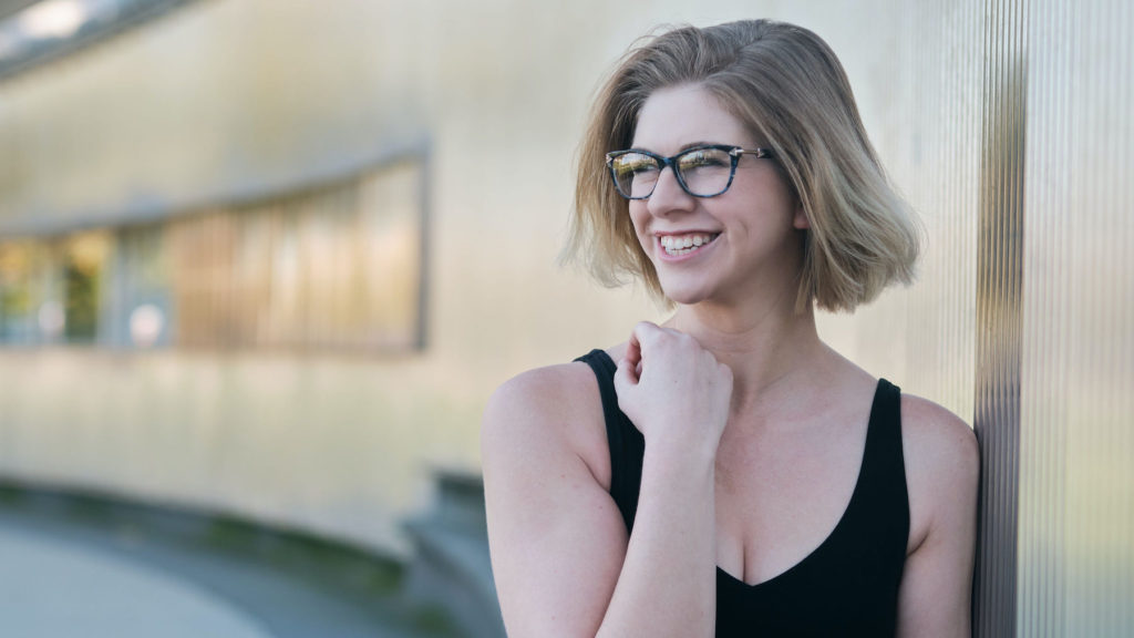 blonde girl with short hair and glasses, wearing a black tank top, looking to the side and smiling in front of an exterior wall