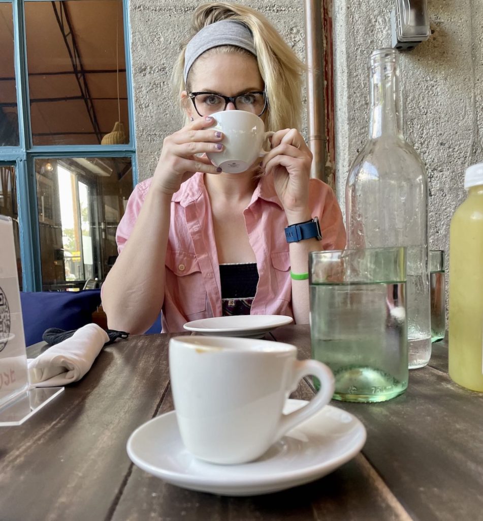 blonde woman with short hair and glasses looking over a white cup of tea, gazing directly at the camera while wearing a blue Fitbit and a pink overskirt. Sitting outdoors at The Rose Venice