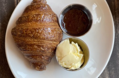 flakey croissant with butter and jelly on the side, resting on a white dish atop a wooden table