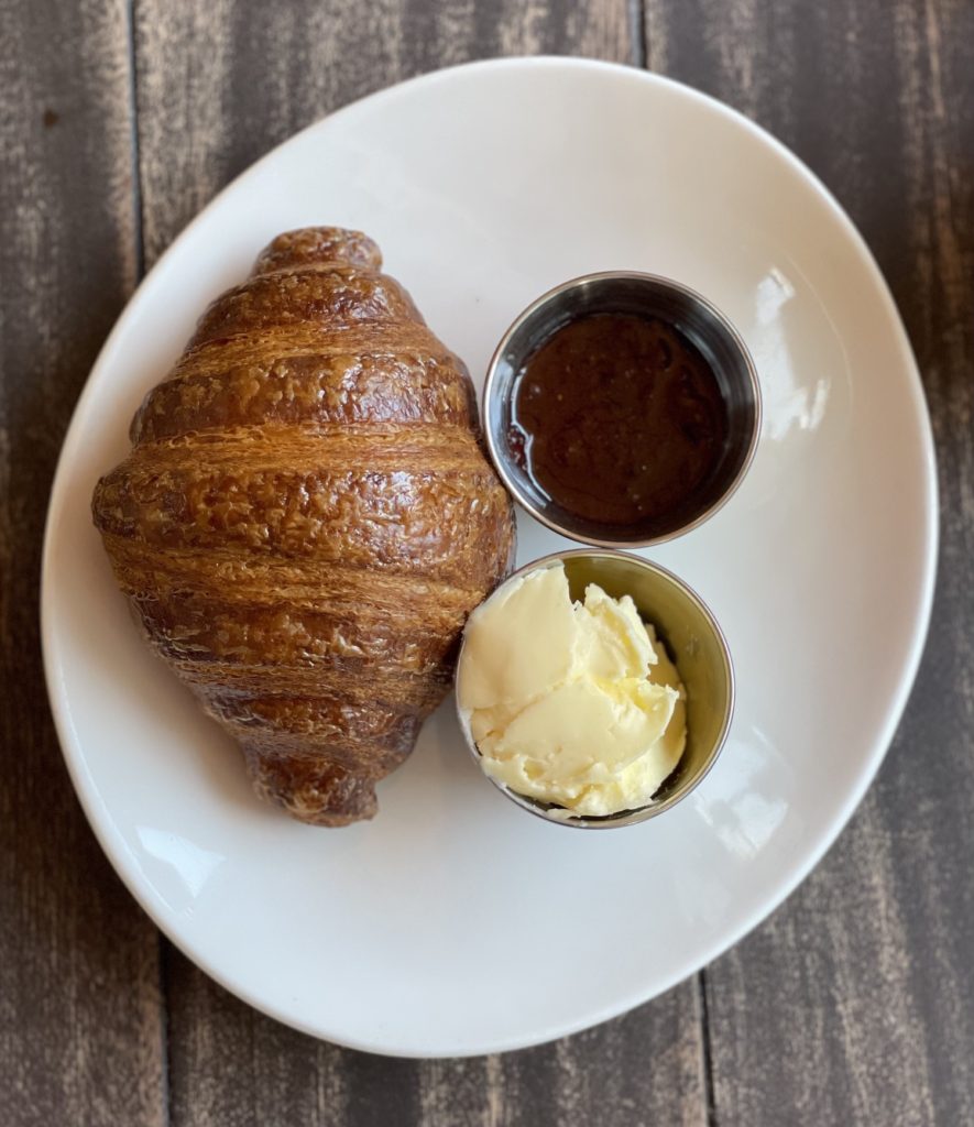 flakey croissant with butter and jelly on the side, resting on a white dish atop a wooden table