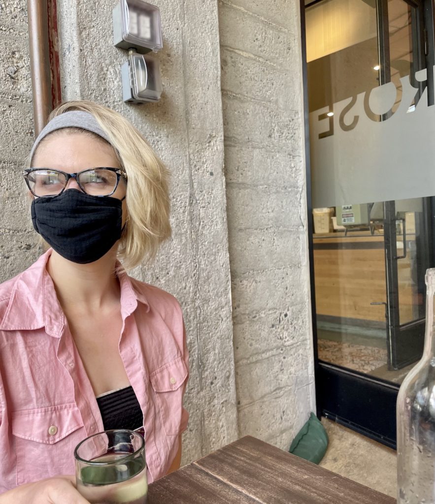 blonde girl with short hair and glasses on looks off on the side, she is wearing a black mask and a pink overskirt, the word "Rose" is visible on the glass door in the background