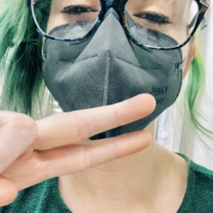 closeup selfie of a girl with a mask and glasses, green hair and a green dress, with her eyes almost closed and showing a peace sign
