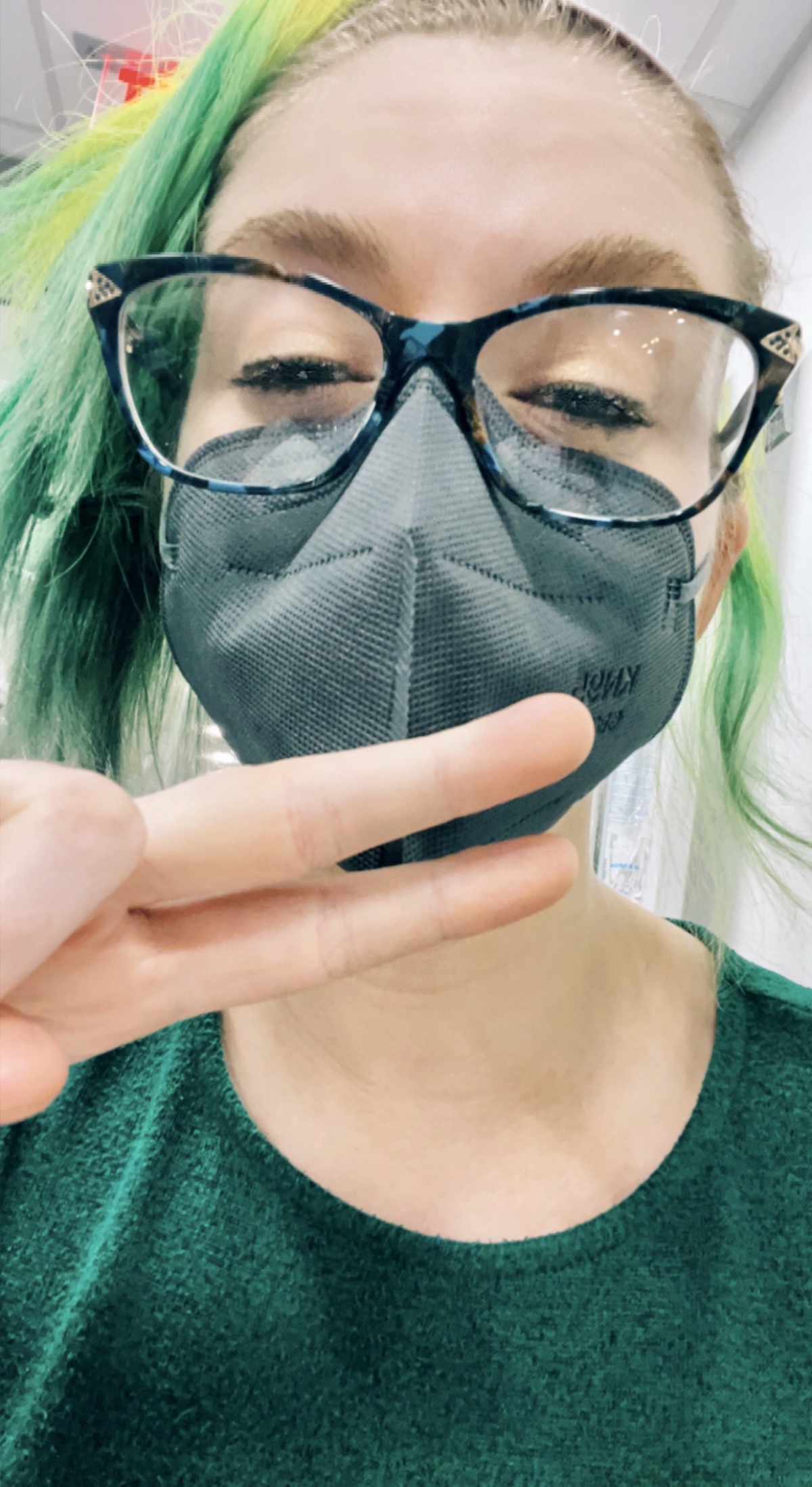 closeup selfie of a girl with a mask and glasses, green hair and a green dress, with her eyes almost closed and showing a peace sign