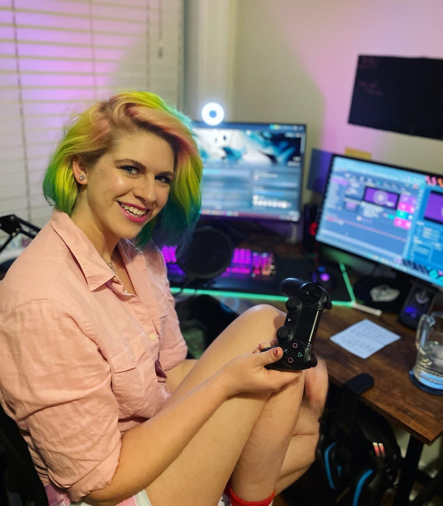 Girl with rainbow colored short hair sitting in a desk chair in front of two computer monitors, wearing a pink long sleeve shirt, holding a PlayStation controller and smiling at the camera