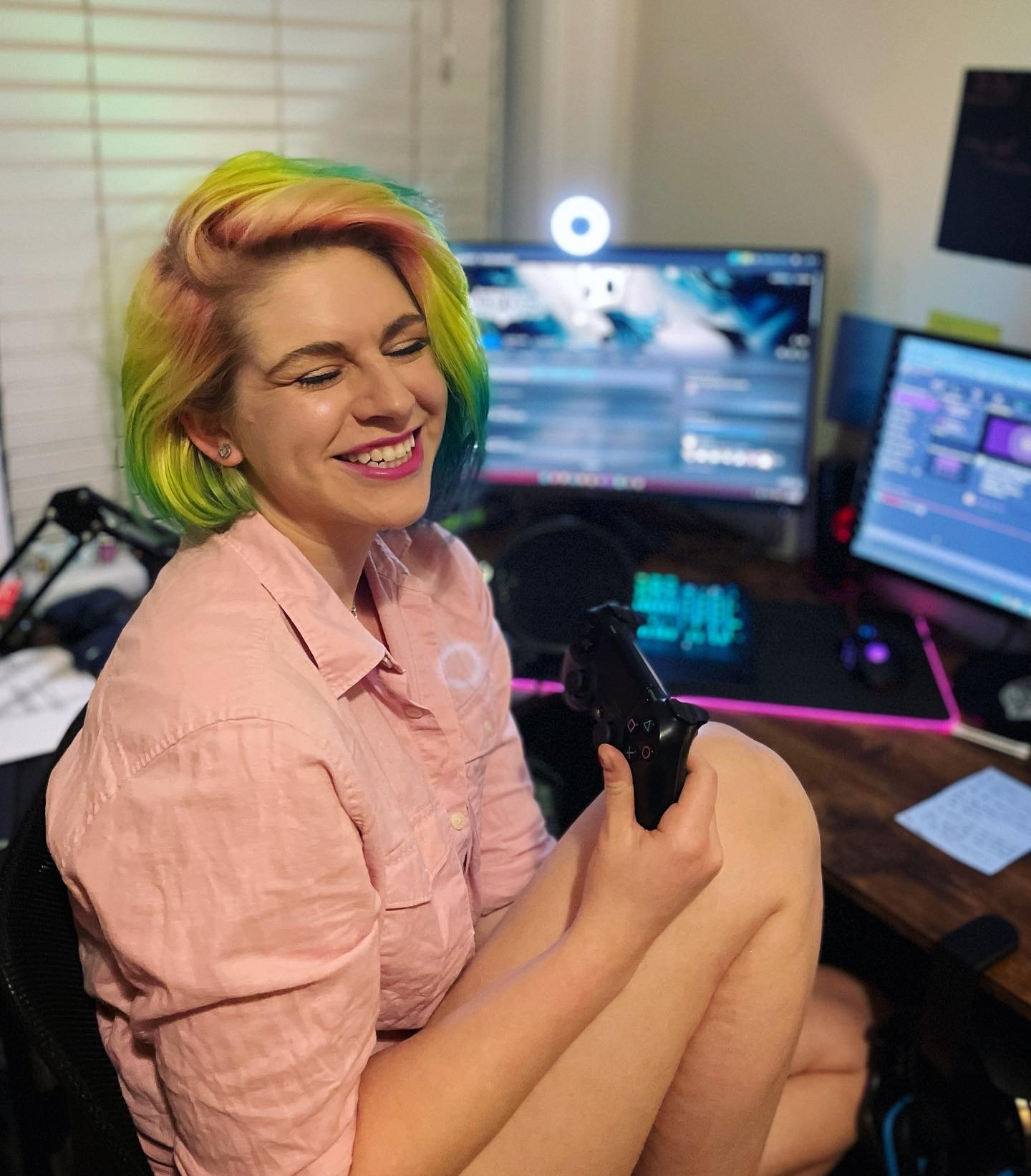 Girl with rainbow colored short hair sitting in a desk chair in front of two computer monitors, wearing a pink long sleeve shirt, holding a PlayStation controller and smiling with her eyes shut