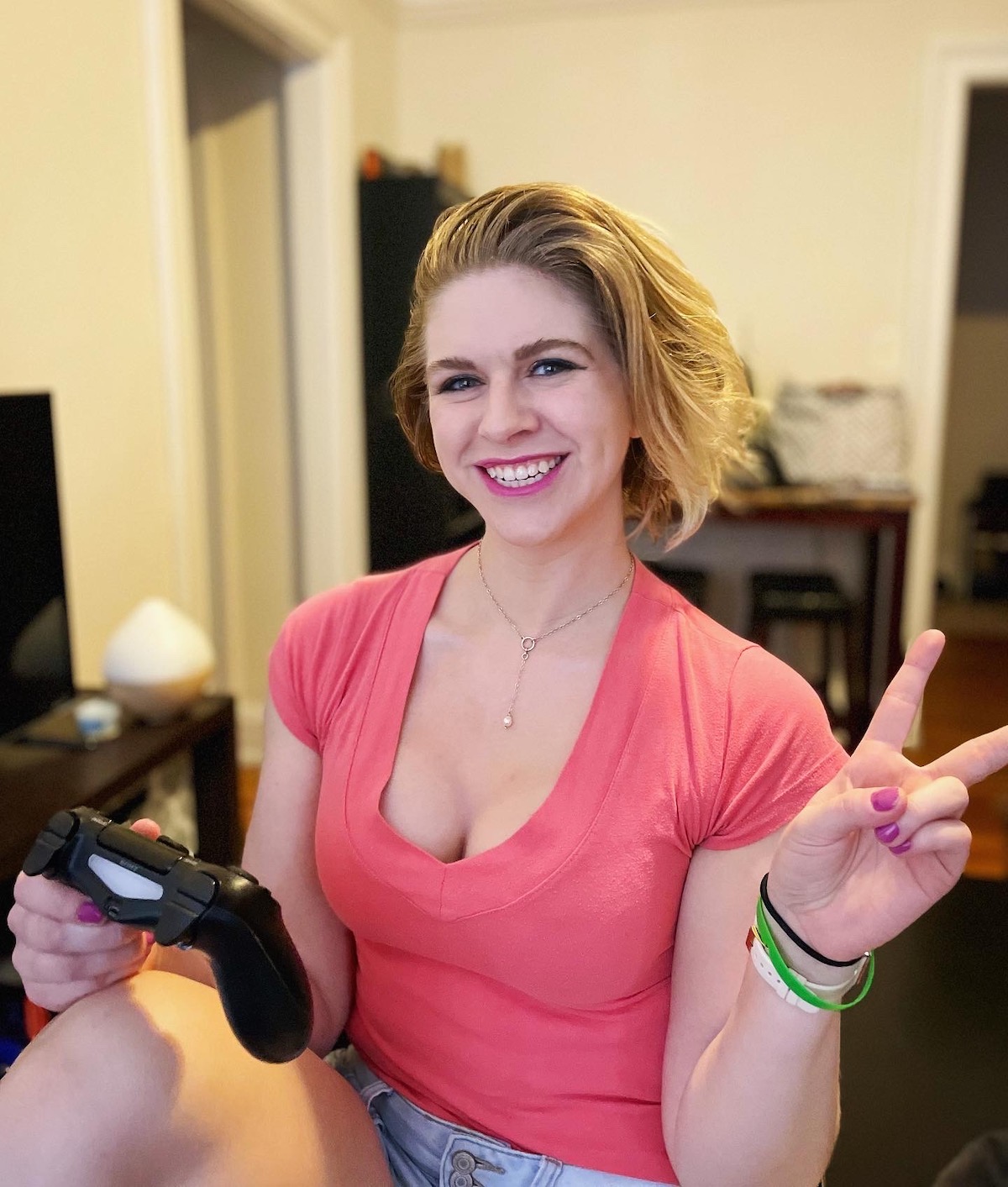 blonde girl with pink lipstick and a peach colored shirt holding a Playstation controller and smiling at camera while throwing the peace sign