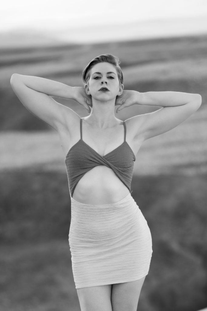 black and white image, model with short cropped hair standing with arms behind her head in front of a blurry exterior background